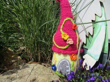 Gehäkeltes Amigurumi-Haus mit pinkem Spitzdach, gelbem Sockel und gehäkelten Blumen, steht auf Stein neben violetten Blüten