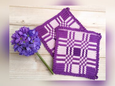 Zwei gehäkelte quadratische Topflappen in Lila und Rosa mit Karo‑Muster neben einer violetten Dekoblume auf Holzuntergrund.