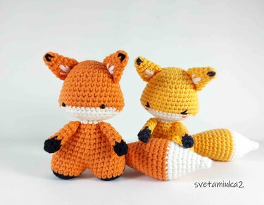 Two crocheted amigurumi foxes, orange and yellow, with white facial markings and dark paws.