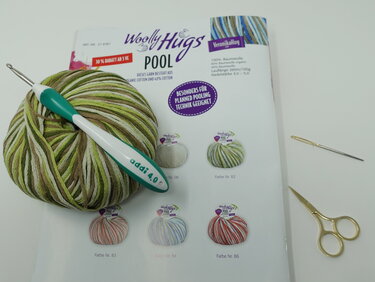 Grün-braun meliertes Garn, ergonomische Häkelnadel, Magazinseite mit Garnmustern und Häkelzubehör auf weißem Untergrund.