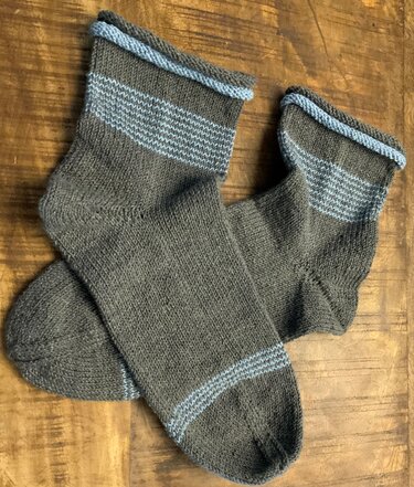Handgestricktes Paar dunkelgrauer Socken mit hellblauen Streifen auf Holzuntergrund.