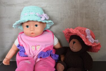 Mintgrüner gehäkelter Hut auf Puppe und korallfarbener gehäkelter Hut auf Teddybär, beide mit Rüschenrand und Schleife.