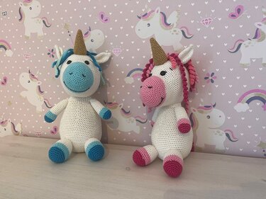 Zwei gehäkelte Amigurumi-Einhörner in Weiß mit goldfarbenen Hörnern, eines mit blauen, das andere mit rosa Details, vor Einhorn-Tapete.
