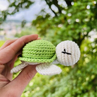 Hand hält einen kleinen gehäkelten Schildkröten-Greifling mit grünem Panzer und weißem Körper vor unscharfem grünem Hintergrund.