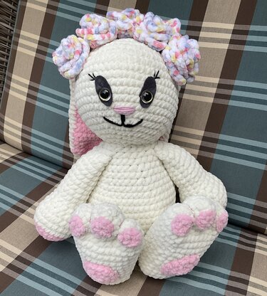 Weißer, gehäkelter Amigurumi-Hase mit rosa Ohren und Pfoten, trägt einen pastellfarbenen Blütenkranz und sitzt auf einem karierten Kissen.