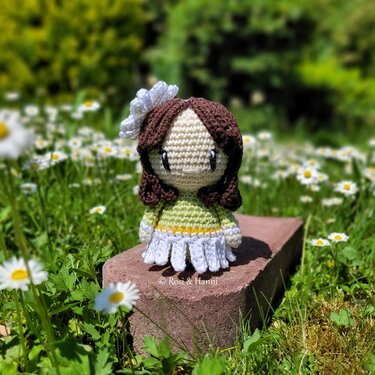 Gehäkelte Amigurumi-Puppe mit braunen Haaren und weißem Rüschenrock auf einem Ziegelstein zwischen Gänseblümchen