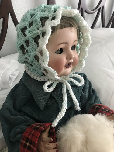 Doll wearing a mint green openwork crochet baby bonnet with white scalloped trim and ties