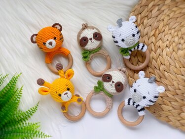 Six crocheted animal head baby rattles on wooden rings arranged on a white faux-fur rug and woven mat