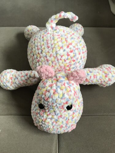 Gehäkeltes, flauschiges Amigurumi-Tier in pastellfarbenen Sprenkeln mit schwarzen Augen und rosa Ohren, liegend auf grauem Stoff