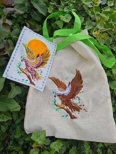 Stofftasche mit braun-weißem Adlerstick, daneben Musterkarte mit gold-rosa Adler und orangefarbener Sonne