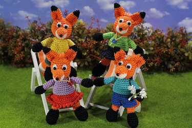 Vier gehäkelte Amigurumi-Füchse in bunten Outfits auf weißen Liegestühlen auf grünem Rasen