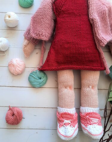 Gestrickte Puppe mit rotem Kleid, rosa Cardigan und Garnknäueln auf weißem Holz