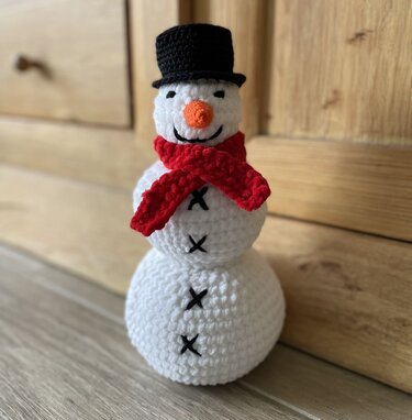 Gehäkelter Amigurumi-Schneemann mit schwarzem Zylinder und rotem Schal vor Holzwand