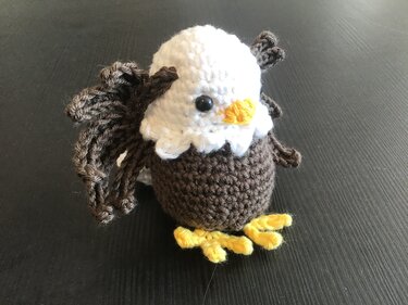 Gehäkelter Amigurumi-Adler mit weißem Kopf, braunem Körper und gelben Füßen auf dunklem Untergrund