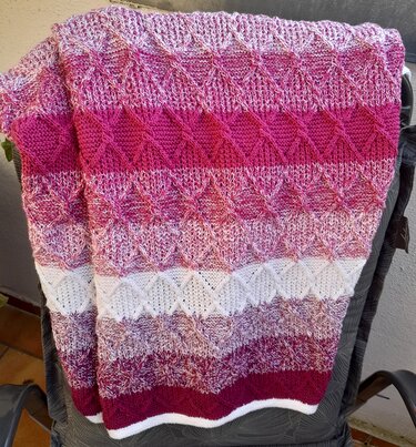 Gestrickte Babydecke mit Rautenmuster in Rosa-, Pink- und Beeretönen, über eine Rückenlehne gelegt.