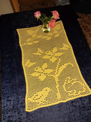 Gelber gehäkelter Filet-Tischläufer mit Vogel- und Blattmotiv auf dunklem Tisch, kleine Vase mit rosa Rosen