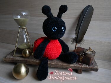 Gehäkelter Marienkäfer in Schwarz und Rot mit blauen Augen sitzt auf einem Holzbrett neben einer Sanduhr und einer Feder.