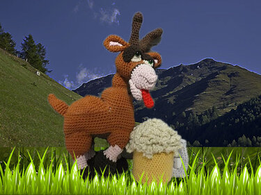 Braunes gehäkeltes Amigurumi-Reh mit grünen Augen, Geweih und roter Zunge neben einem gehäkelten Muffin auf einer grünen Wiese vor Bergen.