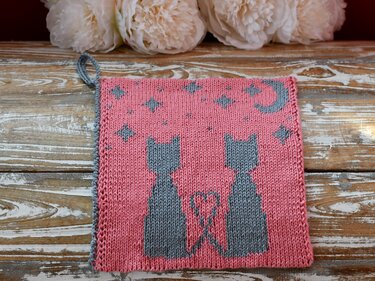 Pink knitted square with two grey cat silhouettes whose tails form a heart, with a crescent moon and stars and a hanging loop.