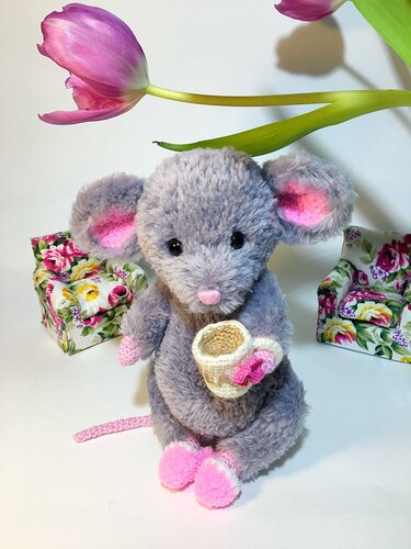 Grey amigurumi mouse with pink ears and feet holding a small crochet cup, floral boxes and pink tulip in background