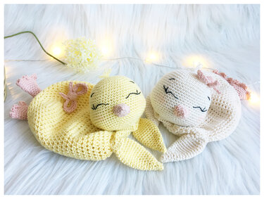 Two crocheted baby loveys — yellow chick and cream bunny with embroidered closed eyes on white faux fur and warm fairy lights