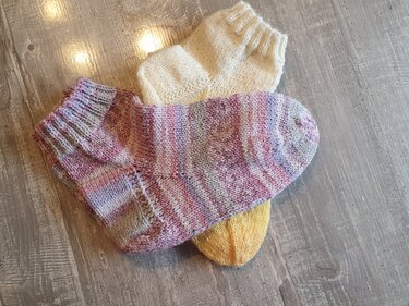 Drei gestrickte Damensocken in Pastell- und Uni-Farben auf hellem Holzboden