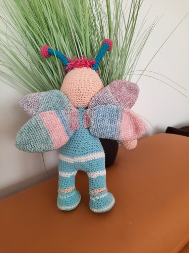Gehäkelte Amigurumi-Schmetterlingsfigur mit pastellfarbenen Flügeln und blau gestreiftem Körper.