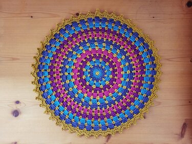 Rundes gehäkeltes Deckchen mit Mandala‑Muster in Blau, Magenta, Lila und Gelb auf hellem Holz
