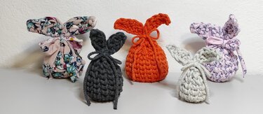Fünf Hasen-Säckchen: drei gehäkelt (dunkelgrau, orange, hellgrau) und zwei genäht aus gemustertem Stoff, jeweils mit Ohren und Bindeband.