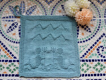 Türkisfarbener gestrickter Topflappen mit Wellen- und Säulenrelief und Aufhängeschlaufe auf bunten Mosaikfliesen