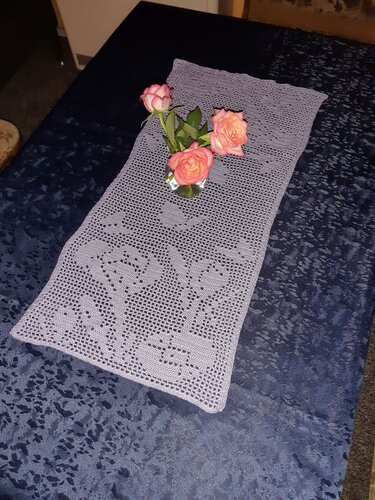 Gehäkelter Tischläufer in Lavendel mit Herzmuster auf dunkelblauem Tischtuch, dekoriert mit drei rosa Rosen in Vase