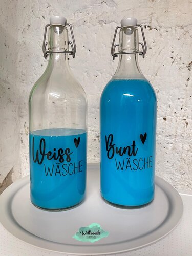 Zwei Glasflaschen mit blauem Inhalt, beschriftet mit schwarzer Plotterschrift: links ",Weiss W
aml;sche
aml;", rechts ",Bunt W
aml;sche
aml;", jeweils mit Herz-Symbol.