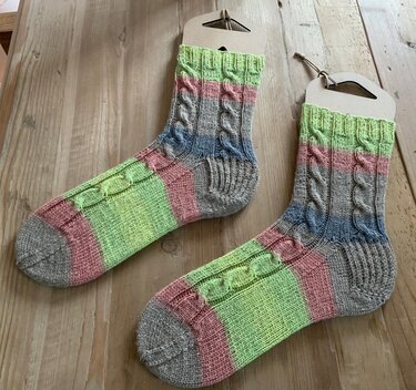Paar handgestrickter Socken mit Zopfmuster und pastellfarbenen Blockstreifen auf Karton‑Sockenform