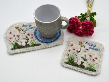 Filzuntersetzer mit gesticktem Hasen, Gras und kleinen pinken Blüten; eine graue Tasse steht auf einem Untersetzer, daneben ein kleiner Blumenstrauß.