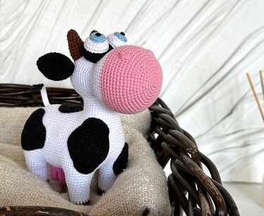 Crocheted amigurumi cow with black spots, large pink snout and blue eyes sitting in a woven basket on beige fabric.