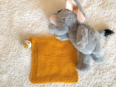 Gestricktes gelbes Babytuch mit Sternstruktur, grauer Plüschelefant und Schnuller auf weißer Flauschdecke
