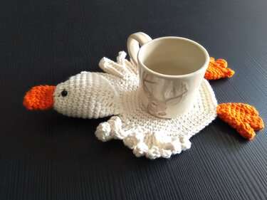 Gehäkelter weißer Gänse-Untersetzer mit orangefarbenem Schnabel und Füßen, darauf eine Tasse mit Hirschmotiv.