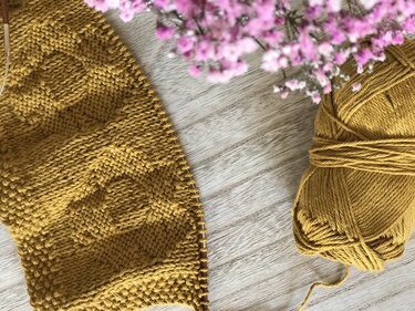 Senfgelbes Strickstück mit Strukturmuster und Stricknadeln, Wollknäuel und rosa Blumen auf hellem Holz.