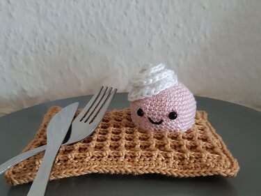 Gehäkeltes rosa Küchlein mit Sahnehaube auf brauner Waffel neben Gabel und Messer