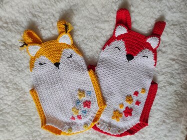 Two crocheted baby fox rompers, one yellow and one red, with white faces, embroidered sleepy eyes, black triangular noses, ear details, floral embroidery, and button closures on a soft white background.