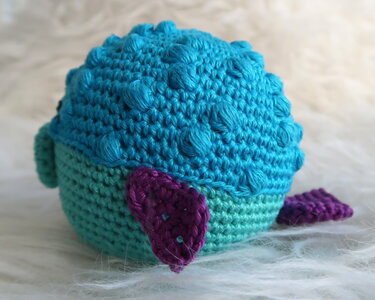 Kugelförmiges blau-türkis gehäkeltes Amigurumi mit Noppenmaschen und lila Flossen