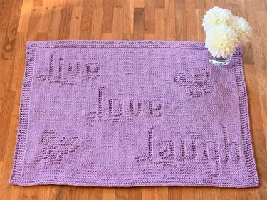 Rectangular purple knitted mat with 'Live Love Laugh' text and heart motifs on a wooden floor beside a small glass of white flowers.