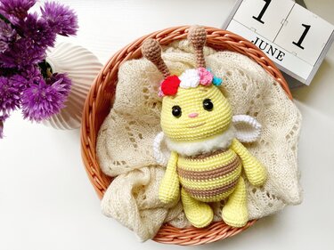 Yellow and brown striped crochet amigurumi bee with multicolored flower crown in a wicker basket on a cream lace wrap.