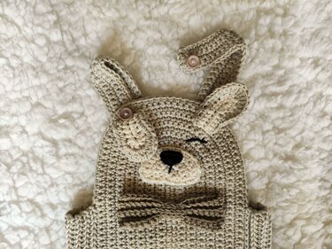 Beige crocheted baby overall with winking dog face, floppy ears and bow tie