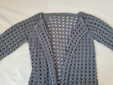 Graue gehäkelte Damenjacke mit langen Ärmeln und durchgehendem Lochmuster, offen ausgebreitet auf hellem Untergrund.