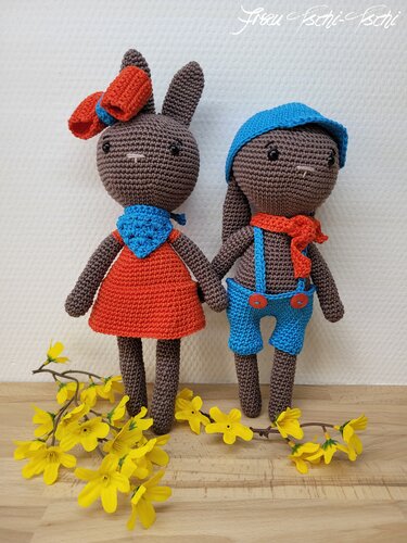 Zwei dunkelgraue Amigurumi-Häkelhasen mit orangefarbenem Kleid und blauer Latzhose, dekoriert mit gelben Frühlingsblüten.