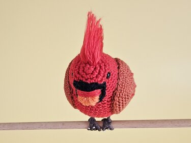 Roter gehäkelter Amigurumi‑Vogel mit aufgestellter Haube auf Holzstange