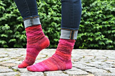 Nahaufnahme von hochgekrempelten Jeans und pink-rot gestrickten Damensocken mit Strukturmuster auf Kopfsteinpflaster vor grüner Hecke.