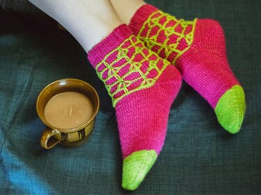 Füße in rosa-grünen gestrickten Socken mit netzartigem Muster neben einer Tasse auf dunklem Stoff
