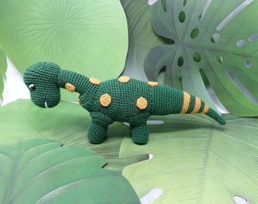 Grüner gehäkelter Amigurumi-Dinosaurier mit gelben Punkten und gestreiftem Schwanz vor Blätter-Hintergrund